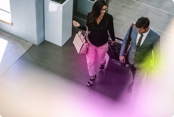 Smiling, bespectacled woman in a black shirt and white trousers entering a modern office wheeling a small suitcase and talking to her male colleague