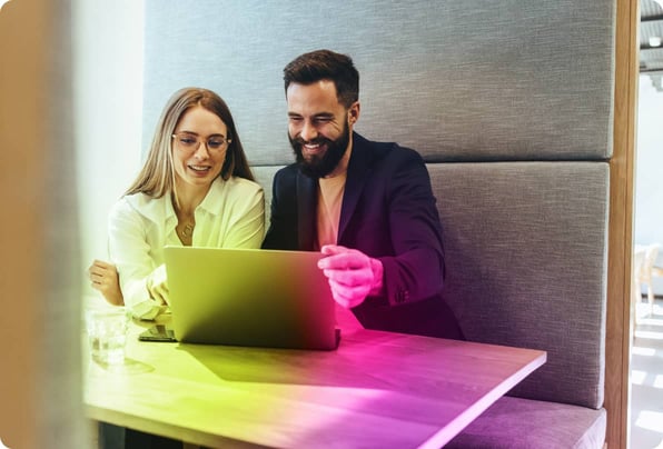 Smiling, bearded man in a t-shirt and blazer sharing his laptop screen with a bespectacled female colleague in a white shirt